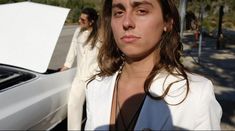 a woman standing next to a white car on the side of the road with another person behind her