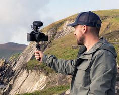 a man holding a video camera on top of a hill next to the ocean with mountains in the background