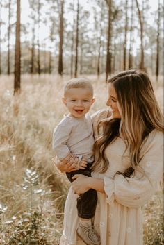 a woman holding a baby in her arms and smiling at the camera while standing in a field