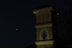 a clock tower lit up at night with the moon in the sky and street lights behind it