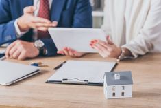 two people sitting at a table with a house model on top of it and papers in front of them