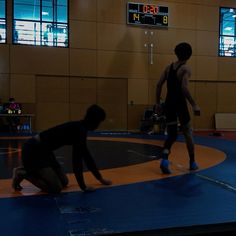 two men are wrestling in an indoor gym with the scoreboard on the wall behind them