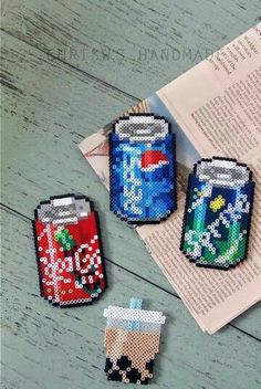 three cross stitch soda cans sitting on top of a table next to an open book