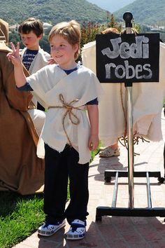 a little boy dressed up as jesus waves to the crowd at an outdoor event with a sign that says jedi robes