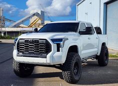 a white truck is parked in front of a blue building with large tires on it