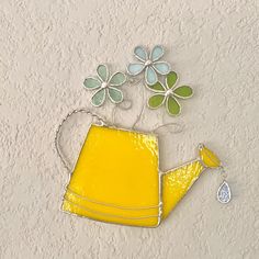 a yellow watering can with flowers in it on a white wall next to a silver chain