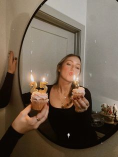 a woman holding two cupcakes in front of a mirror with candles on it