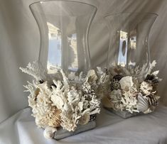 two clear vases with flowers and seashells in them on a white cloth