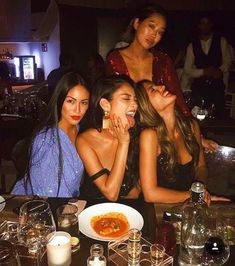 three beautiful women sitting at a table with food and drinks in front of them, posing for the camera