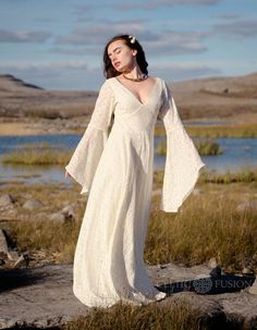 a woman in a long white dress standing on rocks near water and grass with her arms outstretched