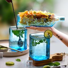 a person holding a glass container filled with water and vegetables on top of a table