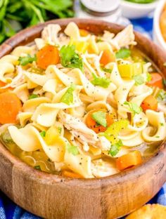 a wooden bowl filled with chicken noodle soup next to crackers and salad greens