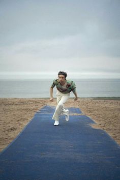 a man in white pants is running down a blue mat on the beach with an ocean in the background