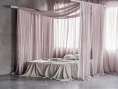 an unmade bed sitting in front of two windows with sheer drapes on them