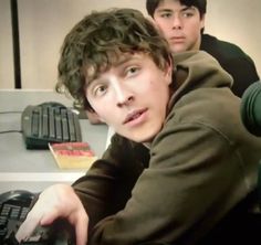 two young men sitting in front of a computer keyboard and monitor, one looking at the camera