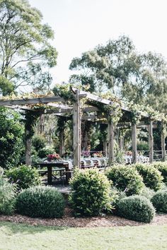 an outdoor dining area is surrounded by greenery and shrubs, with a pergolated arbor in the foreground