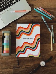 an open notebook sitting on top of a wooden table next to a laptop and pens