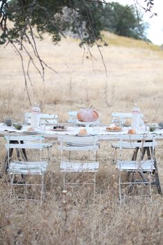 an outdoor table set up in the middle of a field with chairs and plates on it