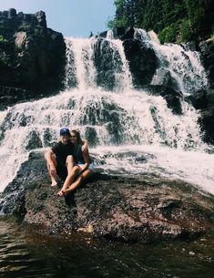 two people sitting on a rock in front of a waterfall