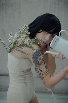 a woman with tattoos holding a white coffee cup and flowers in her hand while standing next to a concrete wall