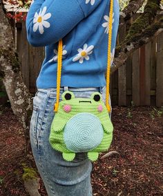 a woman wearing a blue sweater and jeans holding a crocheted frog purse over her shoulder