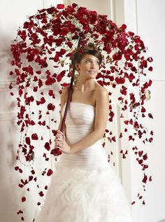 a woman in a white wedding dress holding an umbrella with red flowers hanging from it