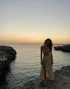 a woman standing on top of a rocky cliff next to the ocean at sun set