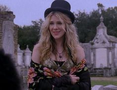 a woman wearing a black hat standing in front of a cemetery with her arms crossed