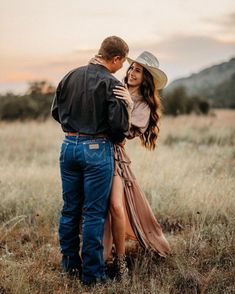 a man and woman standing in the middle of a field with tall grass at sunset