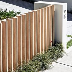 a wooden fence is lined with plants and grass in front of a white box on the sidewalk