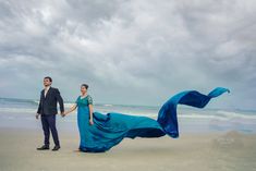 a man and woman holding hands while standing on the beach next to the ocean with their dress blowing in the wind