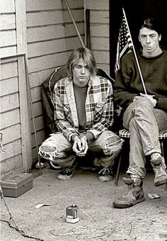 two young men sitting on the porch of a house with an american flag in front of them