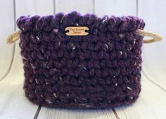 a purple crocheted basket with a wooden tag on the front and bottom, sitting on a white wood surface