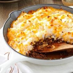 a casserole dish with meat and cheese in it on a white cloth next to a wooden spoon