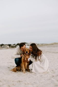 a man and woman kneeling in the sand with their dog