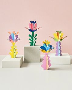 several different colored paper flowers in vases on a white table with a pink wall behind them