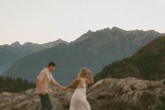 a man and woman holding hands on top of a mountain with mountains in the background