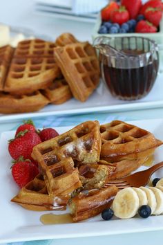 waffles, bananas and syrup are sitting on plates with fruit in the background