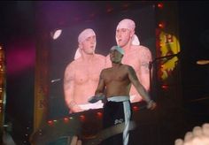 a man with no shirt on standing in front of a large screen displaying two men wearing turbans