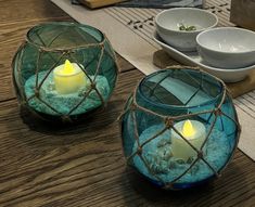 two glass bowls filled with sand and lit candles on top of a wooden table next to plates