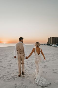 a bride and groom holding hands walking on the beach at sunset in front of hotels