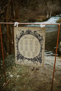 an old - fashioned wedding seating chart hangs from a rusted metal stand in front of a stream