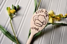 a wooden spoon with the words kiss the cook on it next to some yellow flowers