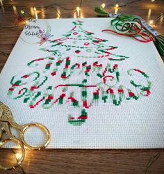 a cross stitch christmas tree on a table next to some scissors and stringed lights