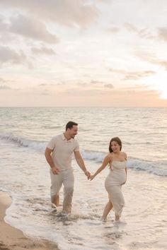 a man and woman holding hands while walking in the water at sunset on the beach