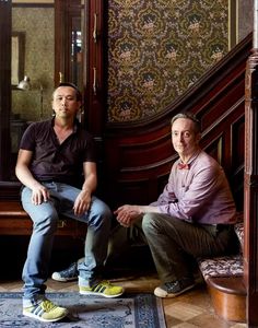 two men sitting on top of a wooden bench in front of a wallpapered room