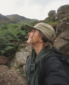 a woman standing on top of a lush green hillside next to a rocky mountain side