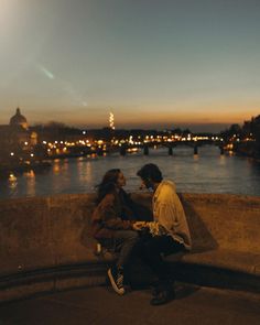 two people are sitting on a ledge near the water at night and one person is looking at his phone