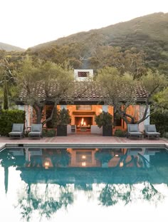 an outdoor fireplace in the middle of a pool surrounded by greenery and trees with chairs around it