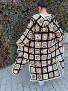 a woman wearing a black and white crochet granny afghan standing in front of a hedge